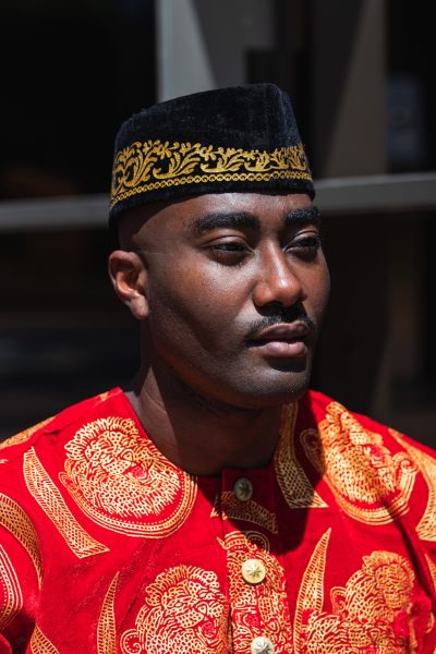 Serious adult African guy in traditional red clothes and kufi cap looking away while sitting in city street near building with glass walls in sunny day
